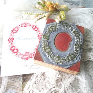 May include: A vintage-style rubber stamp with a floral wreath design, resting on a wooden block. A printed card with the word "Romantiquit&eacute;s" and a floral wreath is next to the stamp. The background includes lace and floral accents.