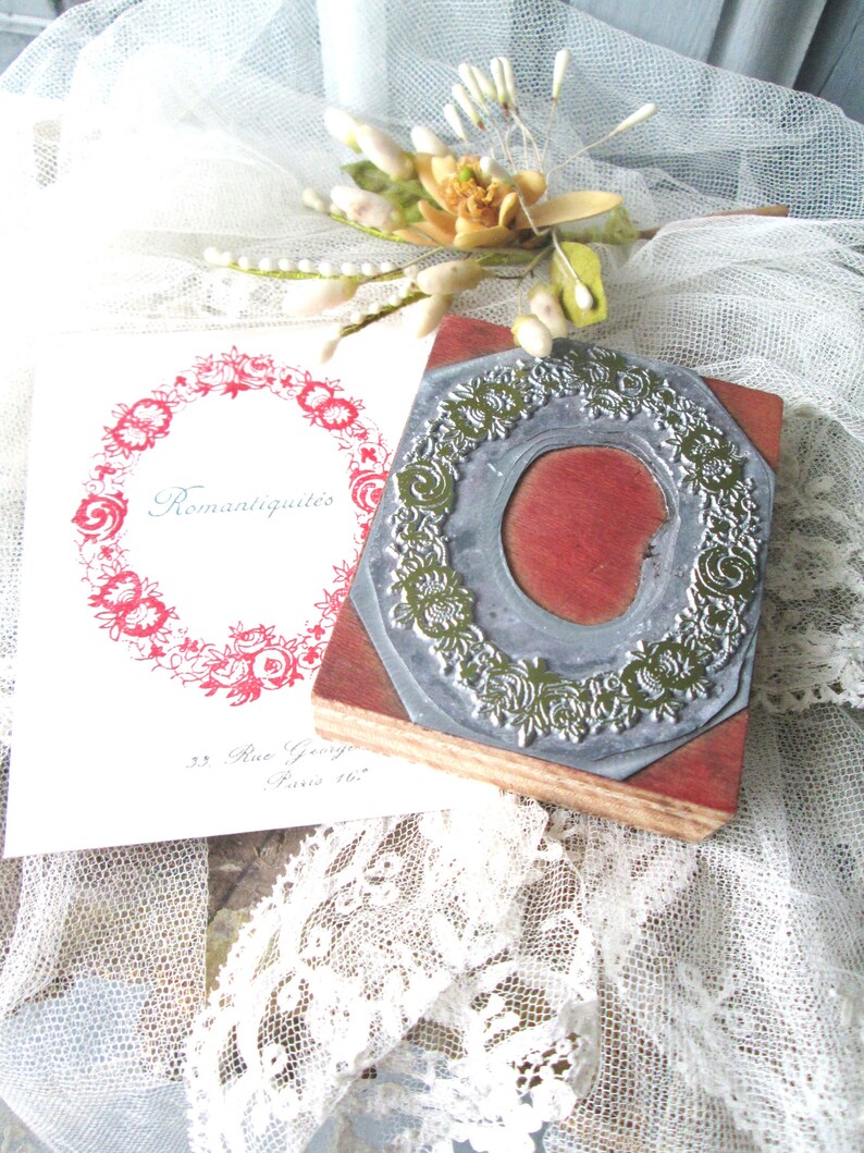 May include: A vintage-style rubber stamp with a floral wreath design, resting on a wooden block. A printed card with the word "Romantiquit&eacute;s" and a floral wreath is next to the stamp. The background includes lace and floral accents.