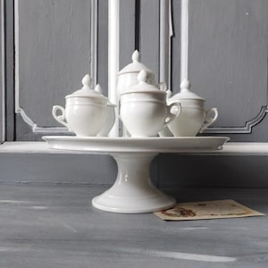 Puede incluir: Un juego de servir de porcelana blanca sobre un soporte para pasteles blanco. El juego incluye cuatro tazas con tapa y un recipiente más grande con tapa. El juego se muestra sobre un fondo gris.