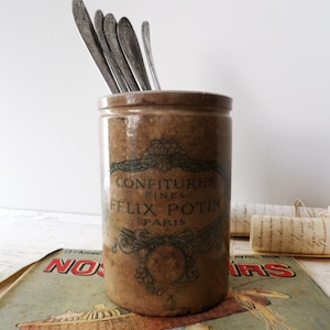May include: A cylindrical brown stoneware crock featuring a dark printed label that reads CONFITURES FINES FELIX POTIN PARIS with decorative scrollwork. Several silver-toned metal spoons are placed inside the container, extending upward.