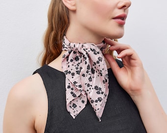May include: A pink neck scarf with a black floral pattern is tied around the neck. The scarf is made of a lightweight fabric and is worn over a black top. The scarf adds a touch of elegance to the outfit.