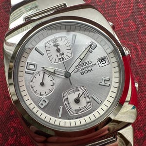 May include: A silver Seiko chronograph watch with a white face and black hands. The watch has a date window at the 3 o'clock position and a small seconds dial at the 9 o'clock position. The watch is on a silver metal band.