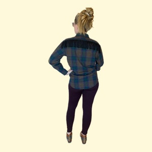 May include: A plaid flannel shirt with black fringe trim on the shoulders. The shirt is worn with dark purple leggings and brown shoes.