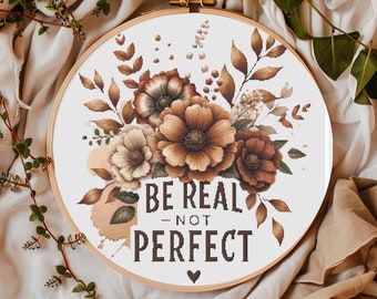 May include: A cross-stitch embroidery hoop with a floral design in brown, beige, and cream. The text "BE REAL - NOT PERFECT" is stitched below the flowers. The hoop is made of wood and has a brass clasp.