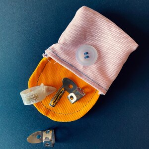 May include: A pink fabric pouch with a button closure, containing a variety of guitar picks and finger picks. The pouch is open and the picks are visible.