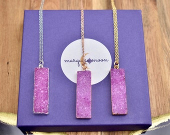 May include: Three necklaces with rectangular, textured, purple pendants. The pendants are set in silver, rose gold, and gold-toned metal. The necklaces are displayed on a purple box with the text "marquise moon" on a white circle.