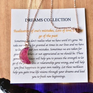 May include: Rose gold necklace with a pink crescent moon pendant. The necklace is displayed on a card with the text "DREAMS COLLECTION" and inspirational text about letting go of the past and finding happiness.