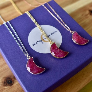May include: Three necklaces featuring crescent moon pendants. The pendants are a deep red hue, framed with gold or silver-coloured metal. They rest on a purple box, with a white circular tag reading "marquismoon". The background is a wooden surface.