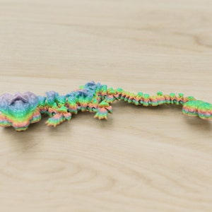 Puede incluir: Un juguete de plástico colorido, segmentado, que se asemeja a un lagarto o un dragón. El juguete está formado por muchas piezas pequeñas que se superponen, cada una con un tono diferente de color arcoíris.