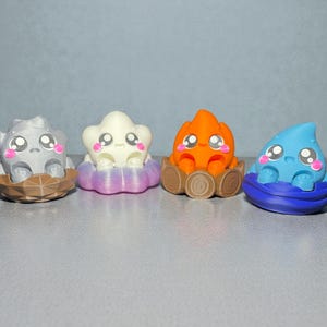 May include: Four colorful, whimsical figurines. A gray rock-like figure, a white star-shaped figure, an orange flame-like figure, and a blue water droplet-shaped figure. Each has large eyes, pink cheeks, and sits in a decorative base.