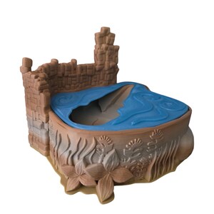 K&ouml;nnte beinhalten: Ein braunes Plastikspielzeugschloss mit einem blauen Plastikpool in der Mitte. Das Schloss hat eine Mauer aus braunen Steinen und einen Turm auf der linken Seite. Der Pool ist von braunen Plastikpflanzen und Blumen umgeben.