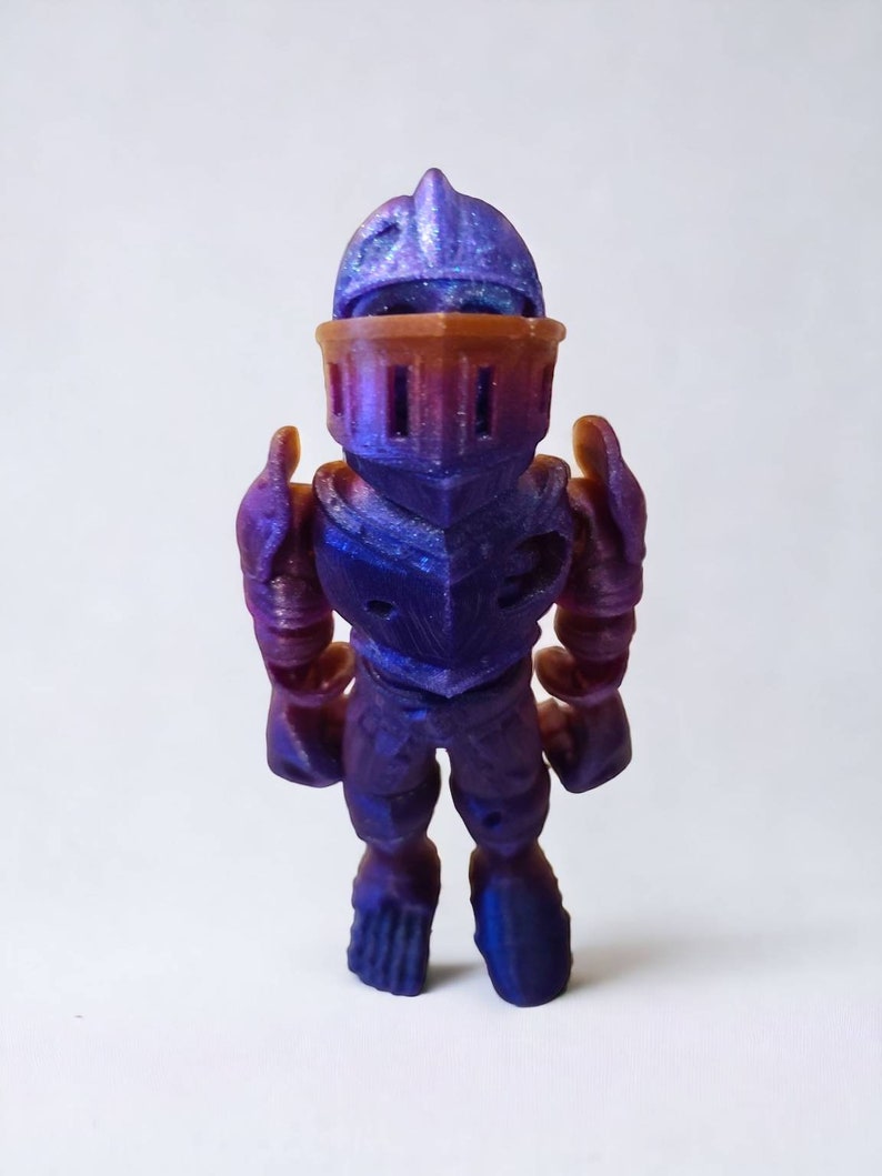 May include: A small, purple and blue plastic toy knight figurine wearing armor. The figurine has a helmet with a pointed top and a detailed face.
