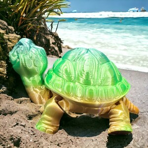 May include: A 3D-printed turtle figurine with a gradient of green, yellow, and blue hues. The turtle is positioned on a sandy beach with the ocean in the background. The figurine has detailed shell and limb textures.