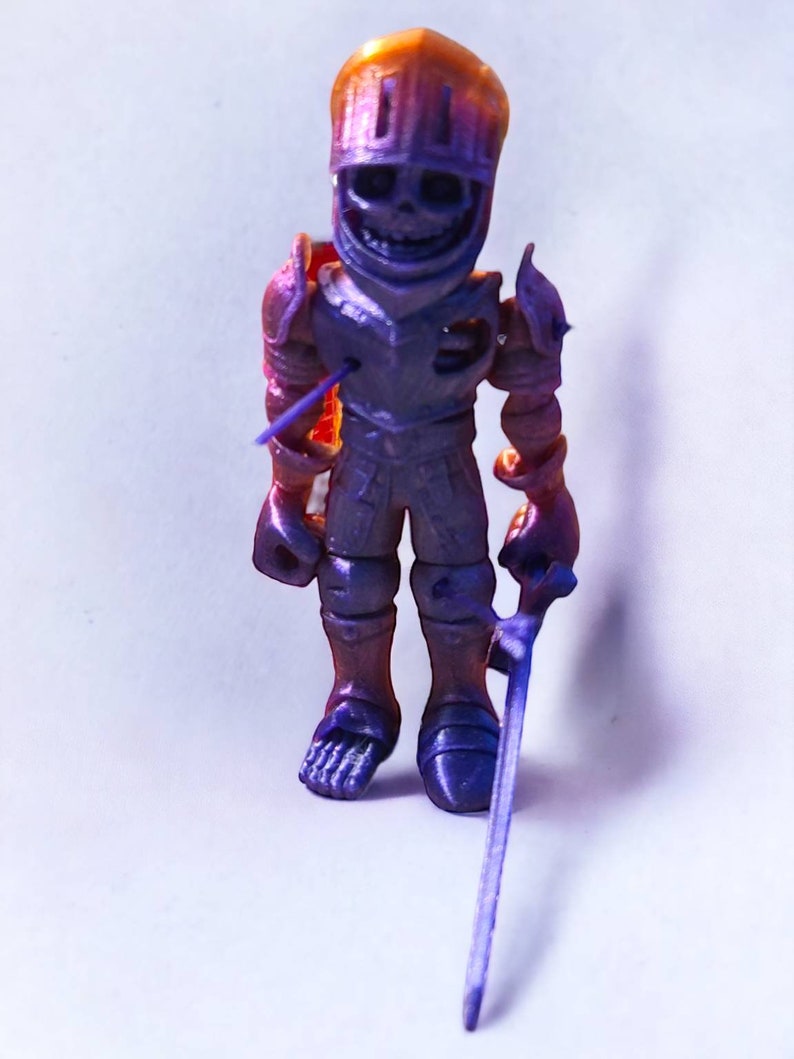 May include: A small, purple and orange plastic toy knight figurine with a sword. The knight has a skeletal face and is wearing armor.