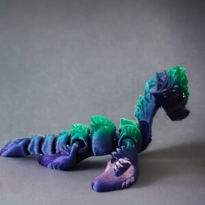 K&ouml;nnte beinhalten: Ein 3D-gedrucktes, artikuliertes, gr&uuml;nes und violettes Seeschlangenspielzeug. Das Spielzeug ist in einer gekr&uuml;mmten Position mit erhobenem Kopf platziert.