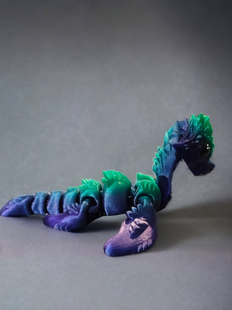K&ouml;nnte beinhalten: Ein 3D-gedrucktes, artikuliertes, gr&uuml;nes und violettes Seeschlangenspielzeug. Das Spielzeug ist in einer gekr&uuml;mmten Position mit erhobenem Kopf platziert.