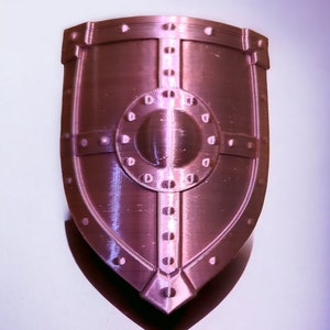 May include: A pink metal shield with a circular design in the center and a raised border. The shield has a textured surface and a pointed bottom.