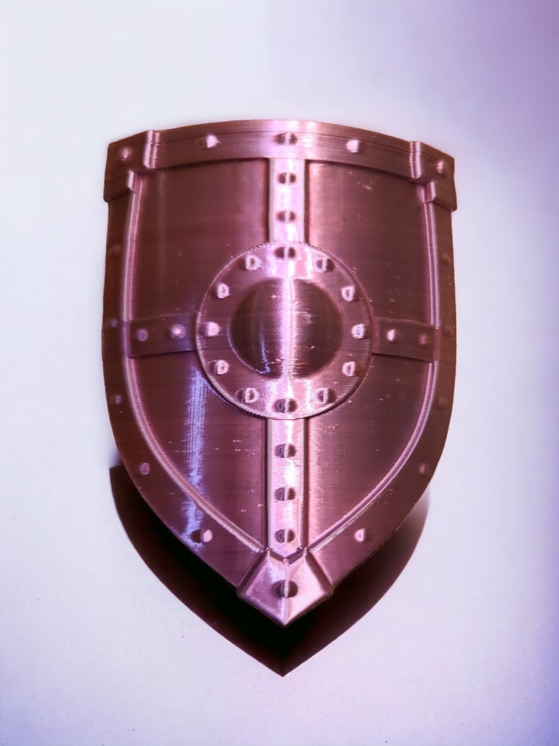 May include: A pink metal shield with a circular design in the center and a raised border. The shield has a textured surface and a pointed bottom.