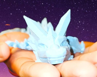 May include: A light blue, crystal-like dragon figurine with a spiky back and a toothy grin, held in a person's hand against a starry night sky.