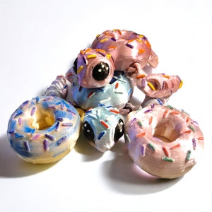 Puede incluir: Una colección de cinco donuts impresos en 3D con coloridos espolvoreados. Los donuts son de varios colores, incluyendo rosa, azul y amarillo. Un donut tiene una cara con ojos grandes.