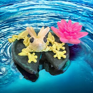 May include: A yellow and white frog figurine sitting on a green lily pad with a pink flower. The frog has large, black eyes and is posed with its legs outstretched.