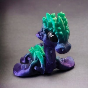 K&ouml;nnte beinhalten: Eine kleine, 3D-gedruckte Drachenfigur mit einem gr&uuml;n-violetten Farbverlauf. Der Drache hat einen stacheligen gr&uuml;nen Kamm und gro&szlig;e schwarze Augen.