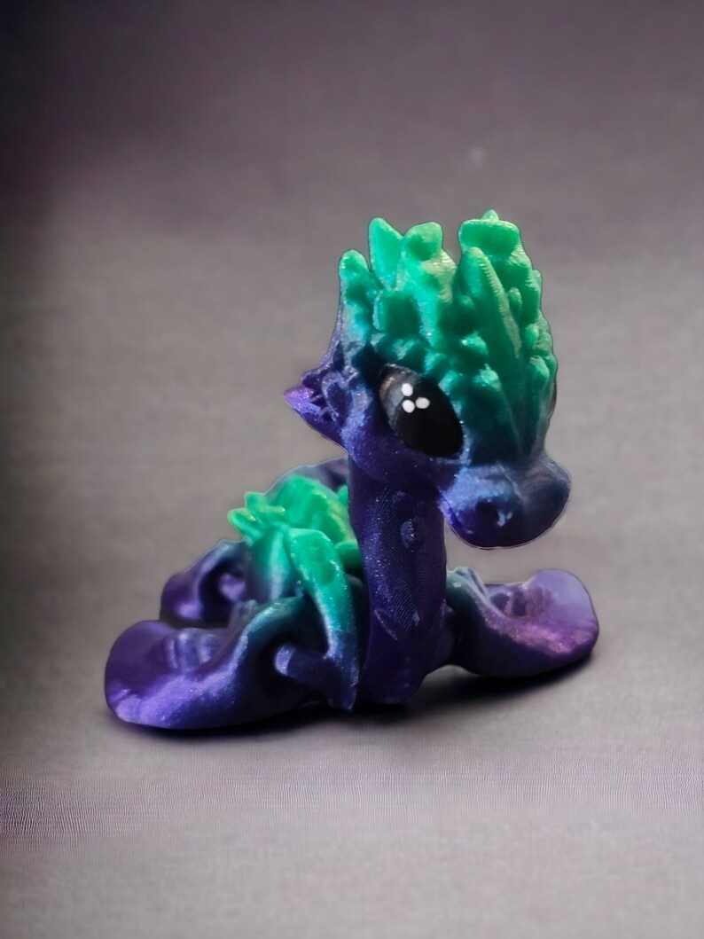 K&ouml;nnte beinhalten: Eine kleine, 3D-gedruckte Drachenfigur mit einem gr&uuml;n-violetten Farbverlauf. Der Drache hat einen stacheligen gr&uuml;nen Kamm und gro&szlig;e schwarze Augen.