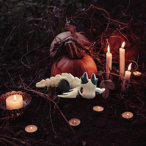 Puede incluir: Una escena de Halloween con una figura de esqueleto de dragón blanco y negro, calabazas, velas encendidas y candelitas. La figura del dragón es el punto focal, rodeada de follaje otoñal, creando un ambiente espeluznante y festivo.