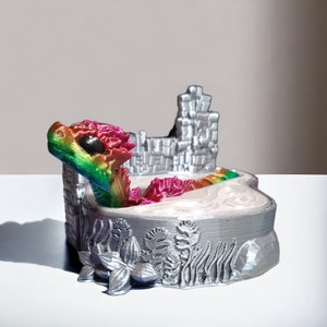 K&ouml;nnte beinhalten: Eine 3D-gedruckte Figur eines Regenbogen-Drachen in einer silbernen Burgkulisse. Der Drache ist in einem Wasserbecken zusammengerollt, hinter ihm eine silberne Burgmauer. Der Sockel der Figur ist mit silbernen Blumen und Pflanzen verziert.