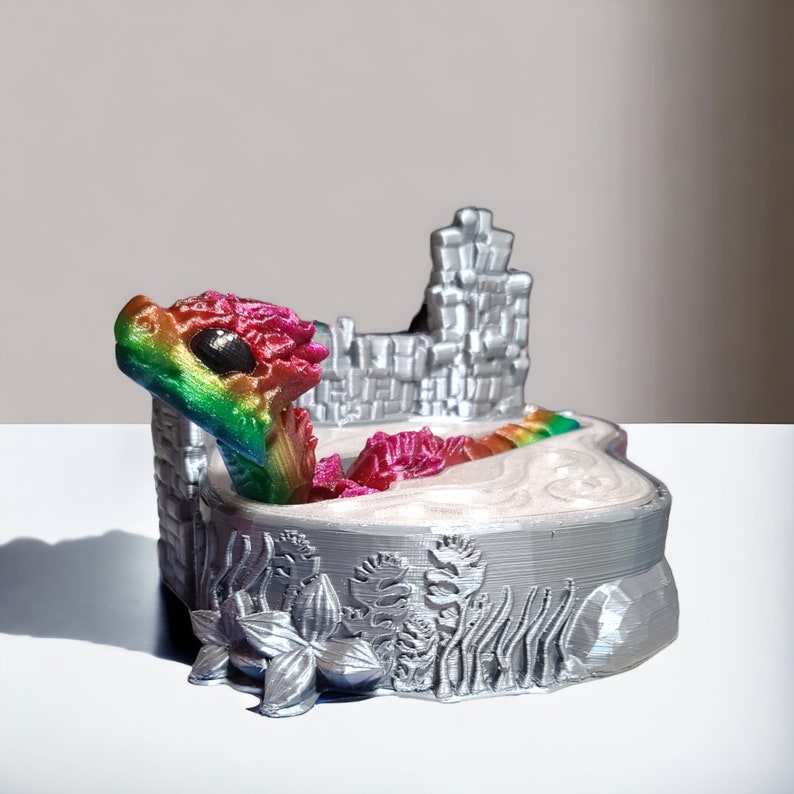 K&ouml;nnte beinhalten: Eine 3D-gedruckte Figur eines Regenbogen-Drachen in einer silbernen Burgkulisse. Der Drache ist in einem Wasserbecken zusammengerollt, hinter ihm eine silberne Burgmauer. Der Sockel der Figur ist mit silbernen Blumen und Pflanzen verziert.
