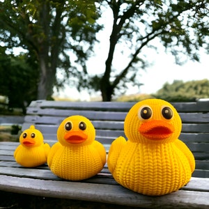May include: Three yellow rubber duck toys of different sizes sitting on a wooden bench. The ducks have knitted textures and orange beaks. The largest duck has large black eyes.