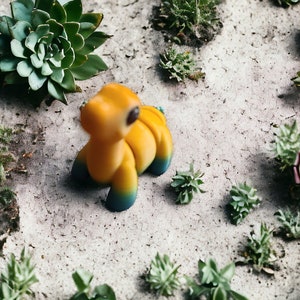 May include: A yellow and blue toy dinosaur figurine with a textured surface. The figurine is sitting on a light gray surface with green succulents.