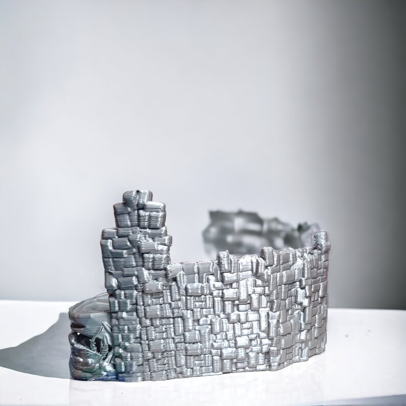 K&ouml;nnte beinhalten: Ein silberfarbenes 3D-gedrucktes Modell einer Ruinenmauer mit einem strukturierten Ziegelmuster. Die Mauer ist gebogen und scheint Teil einer gr&ouml;&szlig;eren Struktur zu sein.