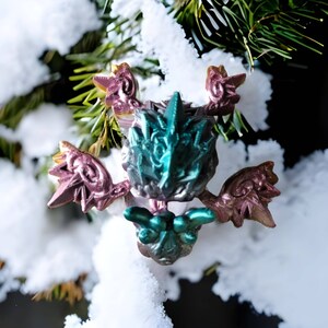 Peut inclure: Une décoration de Noël de couleur turquoise, rose et or en forme de créature mythique ailée. La décoration est ornée de détails complexes et présente un design fantaisiste.
