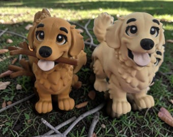 Könnte beinhalten: Zwei Golden Retriever Hundefiguren aus mattem Material. Die linke Figur ist dunkelbeige und hält einen braunen Stock im Maul. Die rechte Figur ist in einem helleren Cremeton gehalten, mit geöffnetem Maul und sichtbarer Zunge.