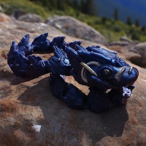 May include: A small, blue, 3D printed dragon figurine with white accents. The dragon is curled up in a defensive position with its head tucked in.