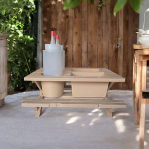 May include: A beige wooden picnic table with two built-in planters. The table has a bench seat and is made of plastic. There are two clear plastic bottles with red caps on the table.
