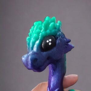 K&ouml;nnte beinhalten: Eine kleine, 3D-gedruckte Drachenfigur mit einem gr&uuml;n-violetten, irisierenden Finish. Der Drache hat einen stacheligen Kamm und gro&szlig;e schwarze Augen.