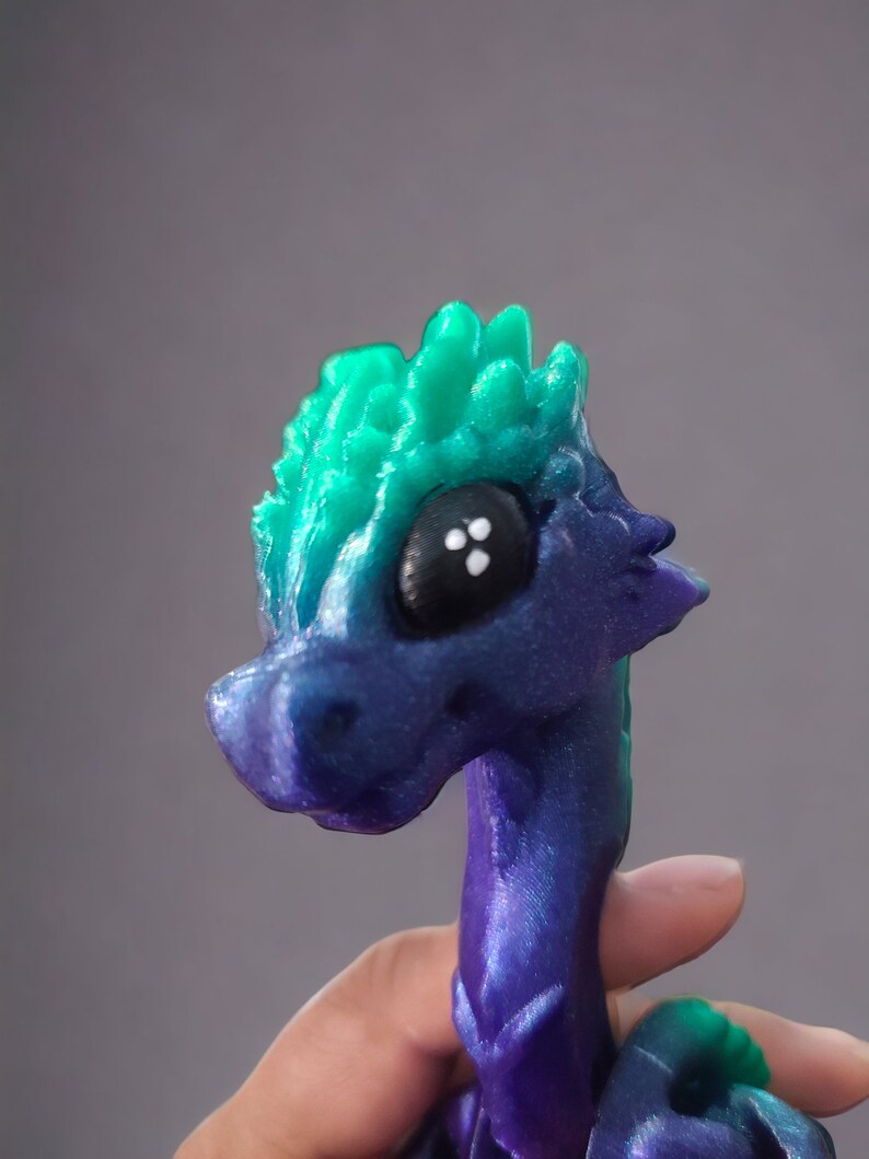 K&ouml;nnte beinhalten: Eine kleine, 3D-gedruckte Drachenfigur mit einem gr&uuml;n-violetten, irisierenden Finish. Der Drache hat einen stacheligen Kamm und gro&szlig;e schwarze Augen.
