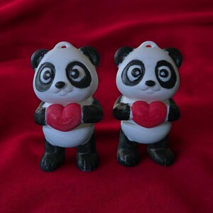 Puede incluir: Dos figuritas de panda blanco y negro sosteniendo corazones rojos. Los pandas están de pie sobre una superficie roja.