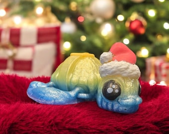 May include: A whimsical, 3D-printed Christmas ornament featuring a snail wearing a Santa hat. The snail's shell is yellow and blue, and the hat is red with white trim. The ornament is set against a blurred Christmas tree background.