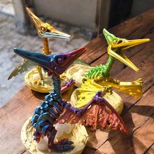 Puede incluir: Tres figuras de dinosaurios esqueléticos de colores en un nido con huevos. Los dinosaurios son iridiscentes con tonos de verde, dorado y morado.