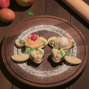 May include: Two turtle-shaped pastries with fruit toppings sit on a wooden board dusted with powdered sugar. Apples, an egg, and a rolling pin are also on the wooden surface. The pastries have tan bodies, green leaf accents, and dark eyes.