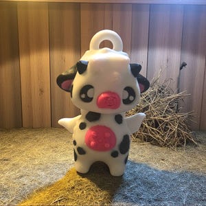 Puede incluir: Figura de vaca blanca con manchas negras, nariz rosa y ombligo rosa. La vaca tiene ojos grandes y expresivos y brazos pequeños. Tiene un lazo en la parte superior para colgar. La figura está en un recinto de madera.