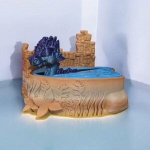 K&ouml;nnte beinhalten: Eine 3D-gedruckte Drachenfigur mit lila und blauen Schuppen sitzt in einem kleinen Wasserbecken. Das Becken ist von einer braunen, 3D-gedruckten Burgmauer umgeben.