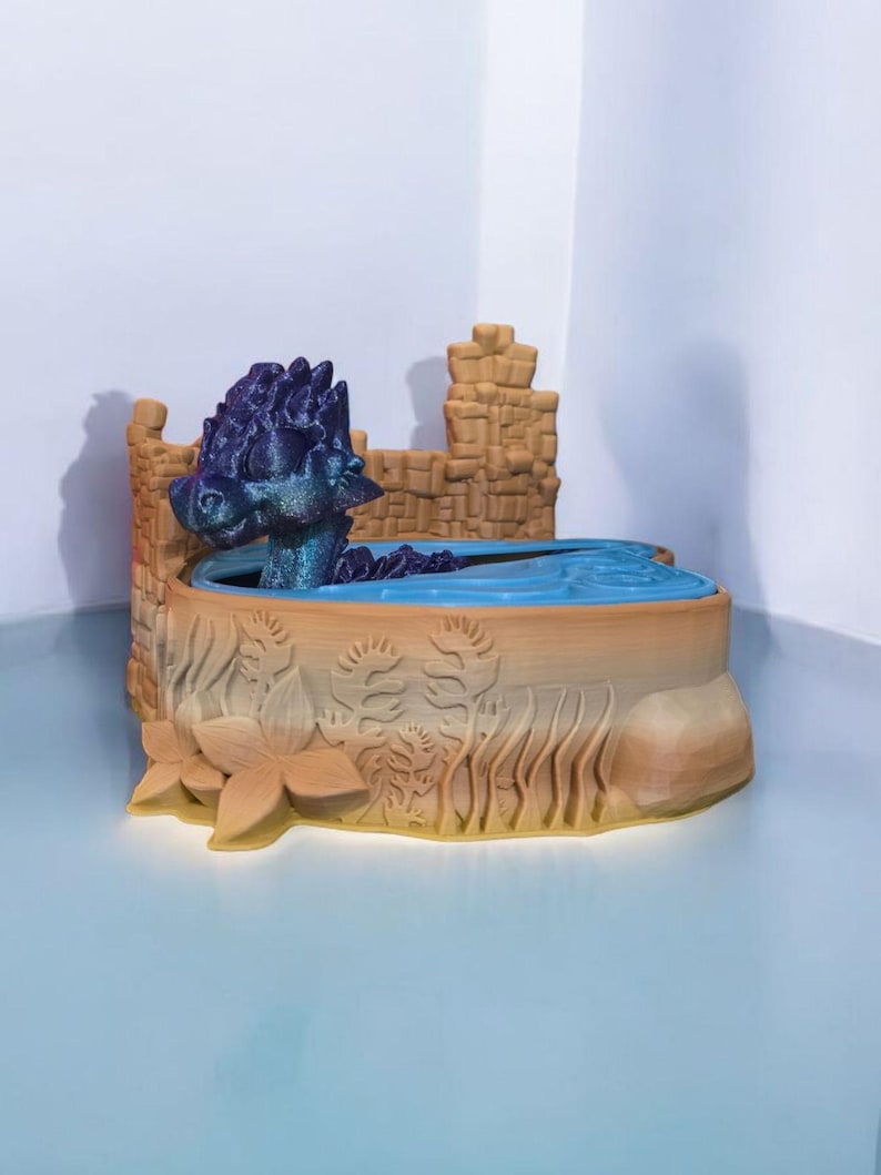 K&ouml;nnte beinhalten: Eine 3D-gedruckte Drachenfigur mit lila und blauen Schuppen sitzt in einem kleinen Wasserbecken. Das Becken ist von einer braunen, 3D-gedruckten Burgmauer umgeben.