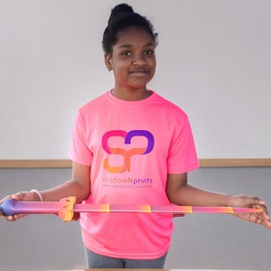 May include: A young person wearing a pink t-shirt with a colorful logo that says "shadowNprints" where prints and ideas collide. They are holding a colorful plastic sword.