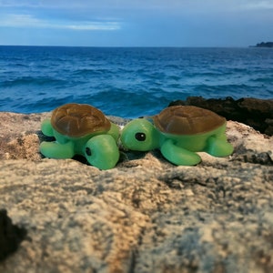 May include: Two small green sea turtle figurines with brown shells, sitting on a rocky surface. The turtles are facing each other, with one slightly tilted to the side.