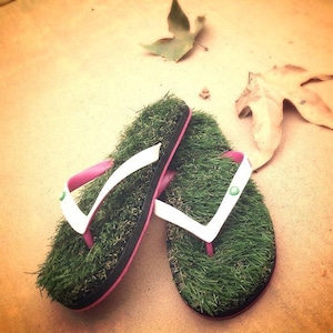 May include: A pair of green grass flip flops with white straps and pink accents. The flip flops are on a light brown surface.