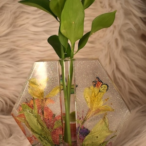 May include: A clear hexagonal vase with a single stem opening. The vase is filled with pressed flowers and butterflies, including a monarch butterfly. The vase has a glittery finish.